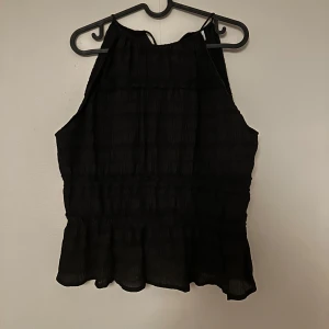 Svart ärmlös blus med volang - Svart blus med hög hals och ärmlös design. Toppen har en lätt rynkad struktur och volang nertill. Helt ny utan prislapp
