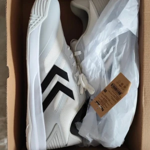 Hummel Dagaz III vita sneakers strl 46 - Snygga Hummel Dagaz III sneakers i vitt med svarta detaljer och grå inslag. Skorna har en modern design med mesh och syntetmaterial, samt en platt sula med coolt mönster för extra grepp. Perfekt för dig som gillar sportig stil och vill sticka ut med klassiska pilar på sidan. Aldrig använda. 