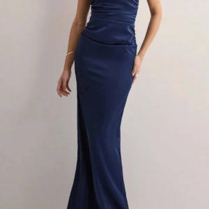 Marinblå satin med tunna straps - Elegant marinblå långklänning i glansigt satintyg med smala axelband som korsas i ryggen. Klänningen har figurnära passform upptill och faller snyggt längs kroppen med slit vid höger sida lår. Perfekt för bal eller festliga tillfällen.  Orginal pris 899kr. 