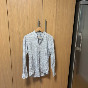 Randig henley-skjorta från Zara - Snygg randig henley-skjorta från Zara i vitt och blått. Skjortan har ståkrage, knappar framtill och långa ärmar med blå armbågslappar. 