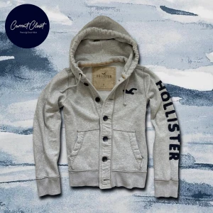 Grå hoodie från Hollister med knappar - Snygg ljusgrå hoodie från Hollister med stora svarta bokstäver på ena ärmen och broderad logga på bröstet | En varm tröja som är perfekt för vintern | Size M | Pris går att diskuteras |