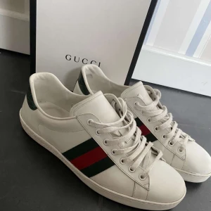 Gucci vita sneakers med röd/grön rand - Säljer ett par klassiska vita sneakers från Gucci i skinn med de ikoniska gröna och röda ränderna på sidorna. Skorna har snörning, rund tå och platt sula. Boxen kommer med på köpet men kvittot finns tyvärr inte. För fler frågor/ bilder skriv pv🤝