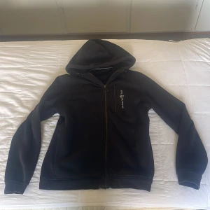 Svart hoodie från Sail Racing med dragkedja - Svart hoodie från Sail Racing med hel dragkedja och broderad logga på bröstet. Tröjan har huva, lång ärm och en bröstficka med dragkedja. Perfekt för dig som gillar sportig och clean stil.