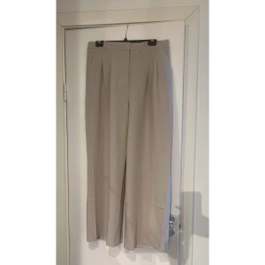 Beiga vida kostymbyxor med pressveck - Snygga beiga kostymbyxor med hög midja och vida ben. Byxorna har pressveck framtill, bälteshällor och fickor i sidorna samt en diskret bakficka. Perfekta för en clean och stilren look. Materialet känns mjukt och har en lätt glans.