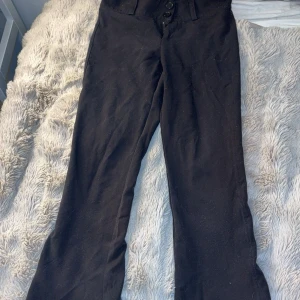 Svarta kostymbyxor från Gina Tricot - Svarta kostymbyxor från Gina Tricot, low waist bootcut. Byxorna har knäppning framtill med flera knappar och bakfickor med lock och knapp. Perfekta till en snygg och stilren look.