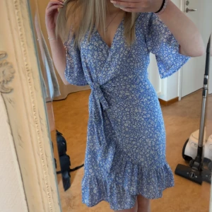 Blå blommig omlottklänning - Supersöt blå omlottklänning med vitt blommönster och volangkant nertill. Klänningen har korta ärmar, v-ringning och knyt i midjan för en snygg passform. Perfekt för sommarens alla tillfällen!