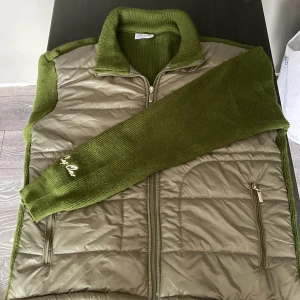 Olivgrön quiltad cardigan All Cap One - Snygg olivgrön dunväst från All Cap One med stickade sidor, rygg och krage. Framsidan är quiltad med dragkedja och två fickor med dragkedja. Broderad logga på ärmen. Perfekt lager-på-lager-plagg med cool mix av material.