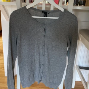 Grå kofta från H&M Basic, storlek M - En enkel och stilren grå kofta från H&M Basic i storlek M. Koftan har rund halsringning, långa ärmar och knäppning framtill med små knappar. Perfekt att slänga över en t-shirt eller linne för en chill look.