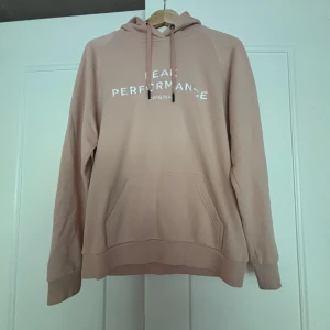 Huvtröja från Peak Performance - Säljer en Rosa-beige hoodie från Peak Performance i storlek L. Tröjan har huva med snörning, stor magficka och tryckt logga framtill. Perfekt för chill dagar eller när du vill ha en enkel och snygg look.