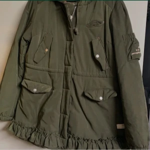 Grön parkajacka Odd Molly med volang - Snygg grön parkajacka från Odd Molly med volangkant nedtill, flera fickor med lock och tryckknappar samt broderad text på ärmen. Jackan har huva och dragsko i midjan för extra passform. Perfekt för dig som gillar unika detaljer och cool stil. Ena sidobfickan är trasig inuti men kan sys om man kan det 