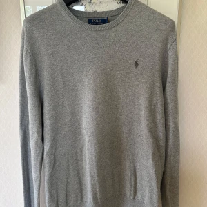 stickad tröja Ralph Lauren - Snygg grå stickad tröja från Polo Ralph Lauren med rund hals och diskret logga broderad på bröstet. Tröjan har långärmad passform och är tillverkad i mjukt material som passar perfekt för kyliga dagar. Enkel och stilren design som funkar till allt.