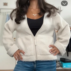 Vit teddyjacka med dragkedja - Mysig vit teddyjacka med hög krage och hel dragkedja framtill. Jackan har två sidofickor och är tillverkad i fluffigt fleece-material som ger en skön och avslappnad vibe. Passformen är normal och ärmarna är långa, perfekt för kyliga dagar.