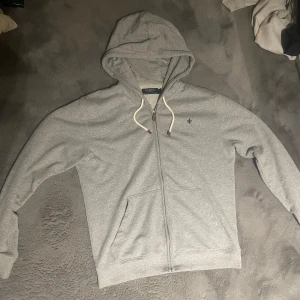 Grå hoodie från Morris med dragkedja - Säljer en stilren grå hoodie från Morris i storlek M. Tröjan har huva med vita snören, dragkedja hela vägen och två fickor framtill. På bröstet finns en snygg broderad logga i mörkblått. Perfekt för chill dagar eller när du vill vara casual. Inga skador, Priset är inte spikat, hör av er vid funderingar ⭐️💫🤩