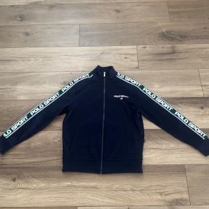 Mörkblå track jacket från Polo Sport - Mörkblå track jacket från Polo Sport Ralph Lauren med hel dragkedja och ståkrage. Snygga vita logotexter längs ärmarna och två fickor framtill. Jackan är gjord i mjuk polyester. Koftan är i barnstolen och skulle motsvara xs. 