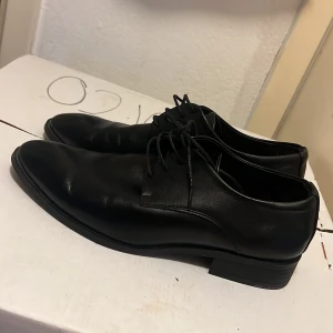 Svarta klassiska skinnskor med snörning - Har använt bara en gång. Snygga svarta skinnskor med klassisk design och spetsig tå. Skorna har låg klack och smidig snörning framtill. Perfekta för en stilren look och passar till både kostym och mer avslappnade outfits.