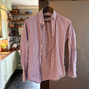 Rutig skjorta från Ralph Lauren, slim fit - Säljer en snygg rutig skjorta från Ralph Lauren i slim fit-modell. Skjortan är lila och vit med klassiskt rutmönster, har långa ärmar och knappar framtill. Ikoniska gröna loggan broderad på bröstet. 