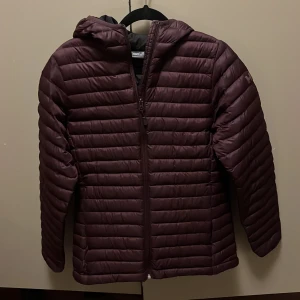 Vinröd dunjacka från Helly Hansen - Säljer en vinröd dunjacka från Helly Hansen med huva och dragkedja. Jackan har en quiltad design och är perfekt för kyligare väder. Den har en normal passform och är tillverkad i syntetmaterial.