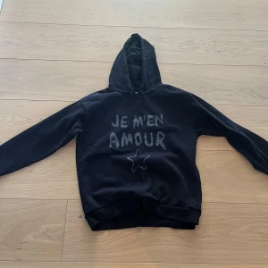 Svart hoodie med text och stjärna - Snygg svart hoodie med texten 'JE M'EN AMOUR' och en stjärna på framsidan. Perfekt för en avslappnad stil med långa ärmar och en klassisk huva.