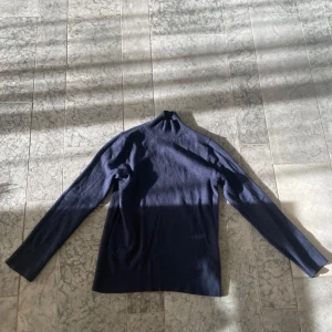 Mörkblå polotröja från Drykorn - Säljer en stilren mörkblå polotröja från Drykorn. Tröjan är ribbad och har långa ärmar, perfekt för kyligare dagar. Den är i storlek M och passar utmärkt för en elegant och avslappnad look. Storlek M för passar bäst för S