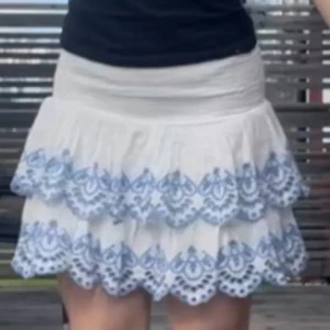 Vit kjol med blå broderi - Charmig vit kjol med vackert blått broderi. Kjolen har en volangdesign och är perfekt för sommarens varma dagar. Den elastiska midjan ger en bekväm passform.