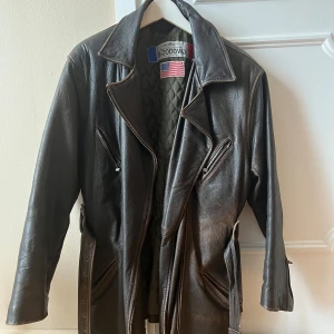 Skinnjacka skinnkappa  - Brun skinnjacka/skinnkappa med bälte i midjan. Jackan har flera fickor med dragkedja och flera innerfickor. Fodrad med ett grönt quiltat foder och ett rutigt foder. Äkta läder, perfekt nu inför våren. Minimala tecken av användning innanför, inget ute