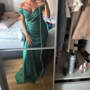 Grön axelbandslös långklänning - Elegant grön långklänning med offshoulder-design och omlott-detaljer. Perfekt för en festlig kväll med sin smickrande passform och vackra drapering. Har dessutom en slits