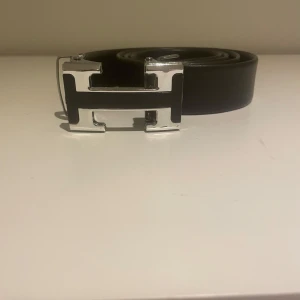 Svart bälte med silverfärgat spänne - Snyggt svart bälte med ett iögonfallande silverfärgat H-format spänne. Perfekt för att ge en stilren touch till din outfit.