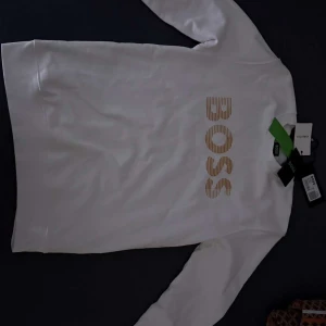 Vit tröja från BOSS - Säljer en stilren vit tröja från BOSS med deras logotyp i guld på framsidan. Tröjan har långa ärmar och en klassisk passform. Perfekt för en avslappnad och trendig look. Tröjan är helt ny bara att skriva för mer info 