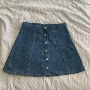 Vintage Jeanskjol med knappar - Snygg jeanskjol med knappar framtill. Perfekt för en avslappnad stil. Kjolen har en klassisk blå denimfärg och en A-linjeform som ger en smickrande passform.
