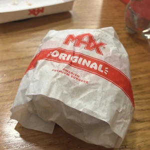 Hamburgare från Max - En hamburgare inslagen i papper från Max, märkt med 'Original' och 'Swedish Premium Burgers'. Förpackningen är vit med röd text och logotyp.