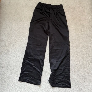 Svarta vida kostymbyxor i satin - Snygga svarta kostymbyxor i satin med hög midja och vida ben. Perfekta för en stilren look. Tillverkade i ett mjukt och bekvämt material som ger en elegant känsla.