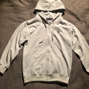Grå hoodie från Polo Ralph Lauren - Säljer en stilren grå hoodie från Polo Ralph Lauren med dragkedja och klassisk logga på bröstet. Perfekt för en avslappnad look. Den har en justerbar huva och fickor framtill.