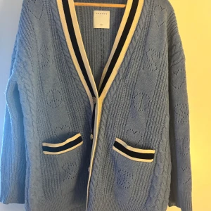 Blå stickad kofta från Sandro - Säljer en snygg blå stickad kofta från Sandro med hjärtmönster och kabelstickade detaljer. Koftan har långa ärmar och två fickor med randiga kanter. Perfekt för kyliga dagar!