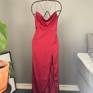 Röd långklänning med korsade axelband i ryggen - Elegant röd långklänning med smala axelband och en slits framtill. Klänningen har en urringning som anpassar sig utefter kroppen. Kommer tyvärr inte till användning längre. Omsorgsfullt använd!