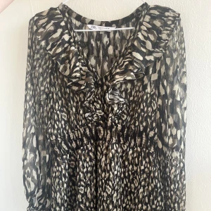 Mönstrad klänning från Zara - Säljer en snygg mönstrad klänning från Zara med volangdetaljer och långa ärmar. Klänningen har ett djärvt mönster i svart och beige och är perfekt för en sommarkväll
