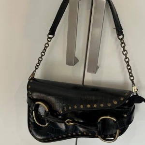 Svart axelväska med nitar - Snygg svart axelväska i skinnimitation med krokodilpräglat mönster. Väskan har guld detaljer och nitar. Inga tecken på användning