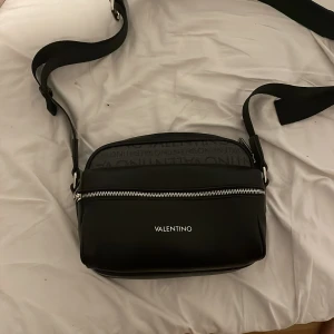 Svart axelväska från Valentino - Snygg svart axelväska från Valentino med justerbar rem och dragkedja. Perfekt för att bära dina nödvändigheter med stil. Väskan har en minimalistisk design med en diskret logotyp på framsidan.