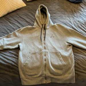 Grå hoodie från Jordan - Säljer en grå hoodie från Jordan med dragkedja och kängurufickor. Den har en klassisk logga på bröstet och en signaturdetalj. Perfekt för en avslappnad stil. MYCKET FINT SKICK stl M