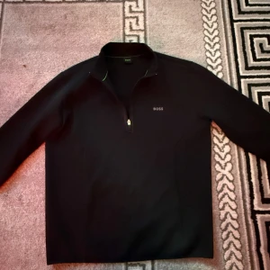 Svart tröja med dragkedja från Boss - Säljer en stilren svart tröja från Boss med dragkedja vid halsen. Tröjan har långa ärmar och en diskret logga på bröstet. Perfekt för en avslappnad men ändå elegant look.