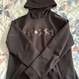  hoodie från Boss - Säljer en stilren svart hoodie från Boss med ett stort logotryck på framsidan. Tröjan har långa ärmar och en klassisk huva. Perfekt för en avslappnad look.