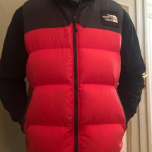 Röd och svart dunväst från The North Face - Säljer en snygg röd och svart dunväst från The North Face. Västen har en dragkedja framtill och är perfekt för kyligare dagar. Den är både stilren och funktionell.