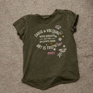 Olivgrön t-shirt från Zadig & Voltaire - Cool olivgrön t-shirt från Zadig & Voltaire med tryck i vitt och rosa. Den har korta ärmar och en avslappnad passform. Perfekt för en avslappnad stil med en touch av attityd.