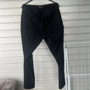 Svarta kostymbyxor från Tailored Originals - Snygga svarta kostymbyxor från Tailored Originals med en klassisk design. Byxorna har knappar och dragkedja framtill samt två bakfickor med knappar. Perfekta för en stilren look.