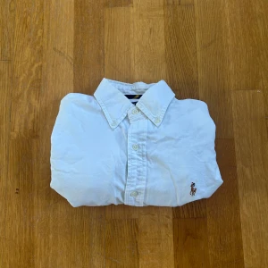 Vit skjorta från Ralph Lauren - Säljer en klassisk vit skjorta från Ralph Lauren med broderad logga på bröstet. Skjortan har knappar framtill och en stilren krage. Perfekt för en stilren look.
