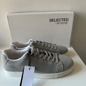 Snygga grå sneakers från Selected Homme i mocka med klassisk snörning. Perfekta för en stilren look. Skorna har en rund tå och vit sula som ger en fräsch kontrast. Helt oanvända!