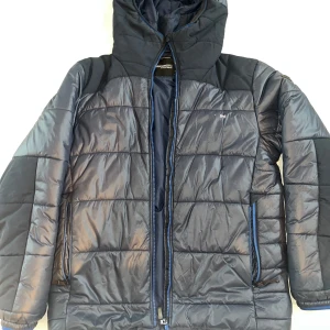 Mörkblå quiltad jacka från Didriksons - Säljer en mörkblå quiltad jacka från Didriksons med huva och dragkedja. Jackan har långa ärmar och är perfekt för kyligare väder. Den har en stilren design med diskreta detaljer och är tillverkad i syntetmaterial. Jackan är i storlek S. 