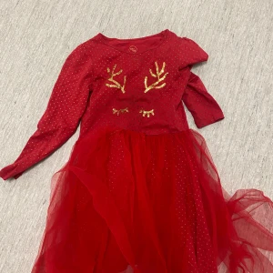 Röd klänning med glitterdetaljer - Säljer en söt röd klänning med glittriga detaljer och renhornsmotiv i guld. Klänningen har långa ärmar och en fluffig tyllkjol som ger en festlig känsla. Perfekt för julens festligheter! 🎄✨