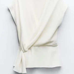 Vit omlottopp - Säljer en stilren vit omlottopp med knytning i sidan. Toppen har en elegant drapering och är ärmlös, vilket gör den perfekt för varmare dagar.