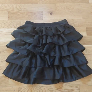 Svart volangkjol  - Snygg svart kortkjol med flera lager av volanger. Perfekt för att ge en extra touch till din outfit. Kjolen har en elastisk midja för bekväm passform. Säljer för den inte kommer till användning längre.