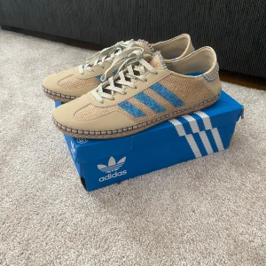 Adidas x Clot Gazelle Linen beige/blå sneakers - Unika Adidas Gazelle Clot sneakers i beige och blått med flätad ovandel och blå detaljer. Snygga blå ränder på sidan, beige skinnpaneler och coola sömmar. Gummisula med Adidas-loggor undertill och snörning framtill. Perfekt för dig som vill sticka ut med stilen. Sällsynta med nypris på 2400kr